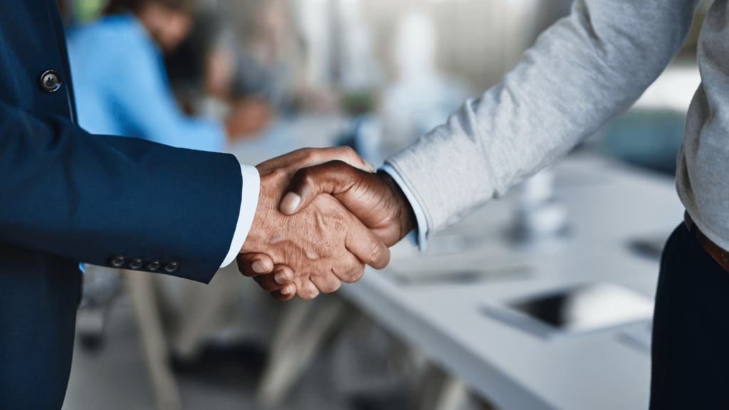 Close-up of two people shaking hands, one in a dark blue suit and the other in a light gray sweater, symbolizing a professional agreement or partnership in a modern office setting.